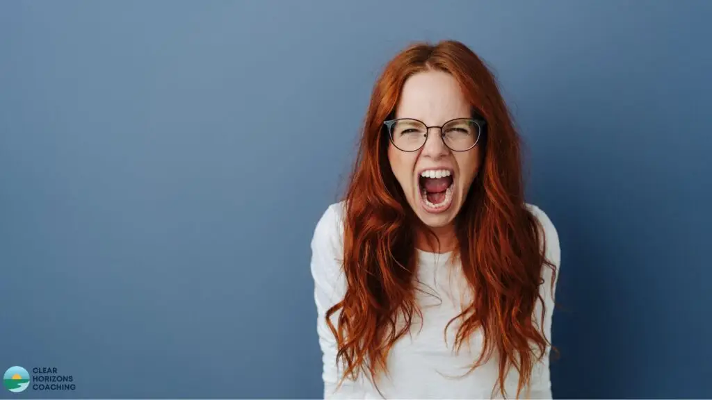 Red-haired woman with glasses is staring straight out of the picture. Her mouth is wide open and she is angry / screaming