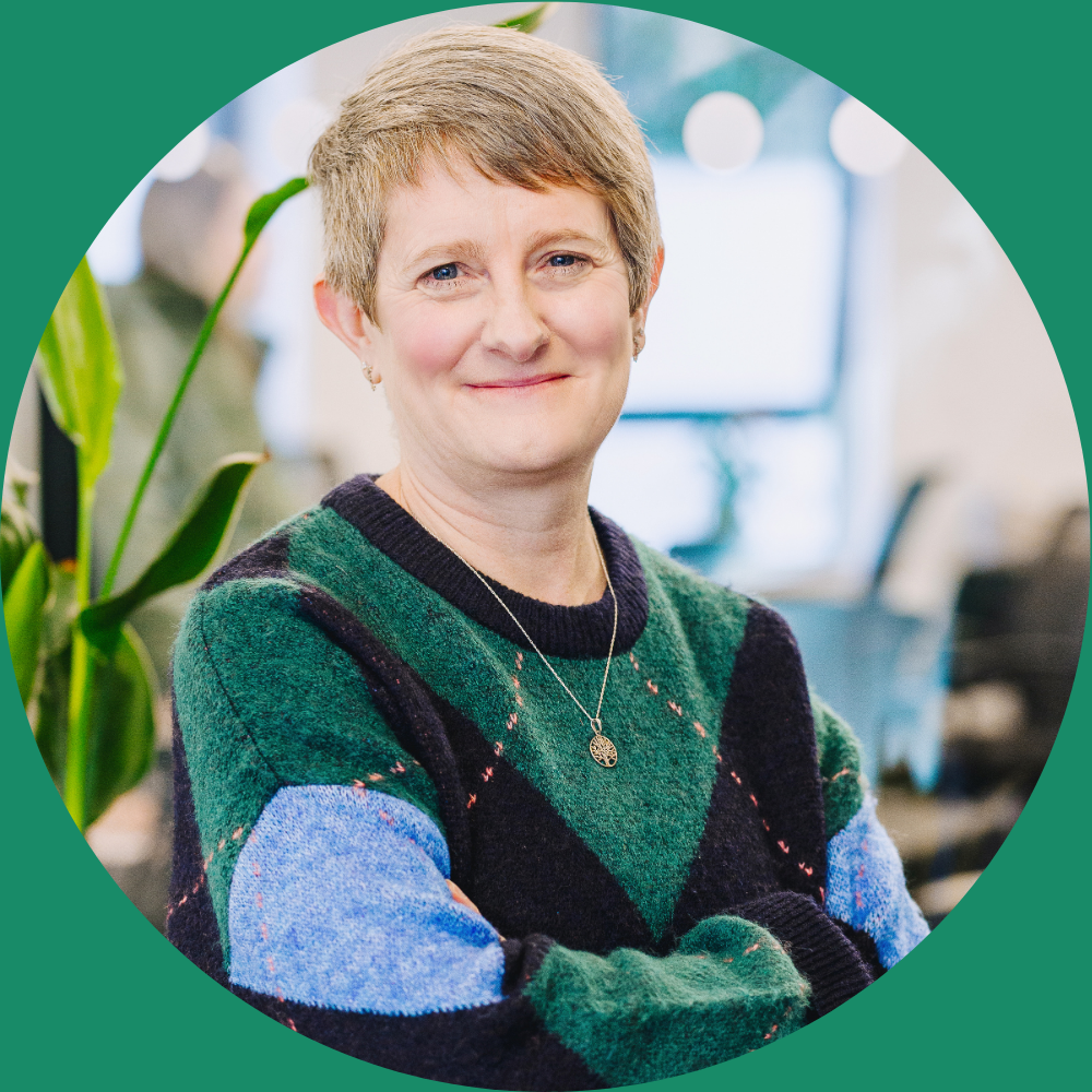 Image shows headshot of Sam standing sideways on to the camera and looking into the lens. Sam is wearing a jumper with blue and green diamonds on it. She has grey hair and is smiling. There are plants in the background in soft-focus