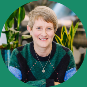 Headshot of Sam, a white woman in her 50s with short grey hair. Sam is looking at the camera and smiling. In the background there are plants and tables in soft focus.