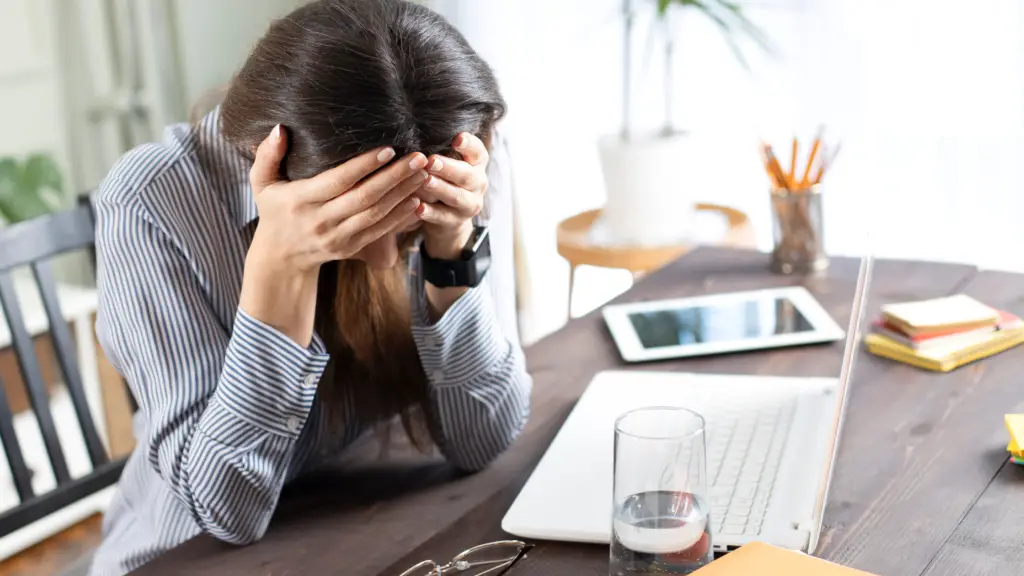 Woman sitting at desk where she has been working as there is a laptop & phone. She has her head in her hands obscuring her face