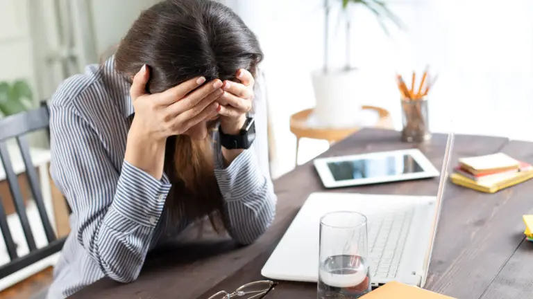 Woman sitting at desk where she has been working as there is a laptop & phone. She has her head in her hands obscuring her face
